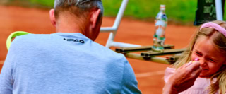 Ein Mann in einem blauen Hemd sitzt auf einem Tennisplatz, w&auml;hrend ein M&auml;dchen mit rosa Haarband l&auml;chelt. Im Hintergrund sind ein Stuhl und eine Wasserflasche zu sehen.