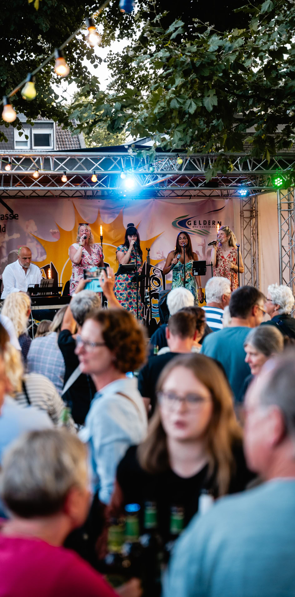 Menschenmenge vor einer B&uuml;hne mit Musikern, umgeben von B&auml;umen und Lichtern.