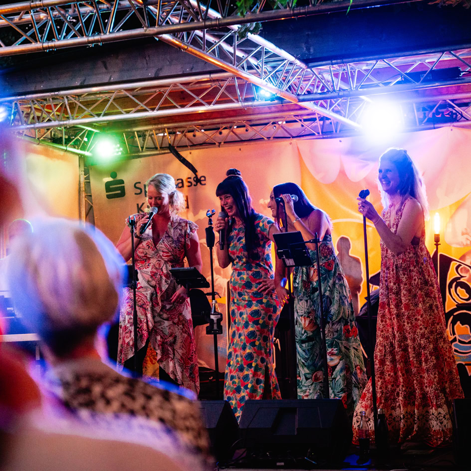 Vier Personen singen auf einer B&uuml;hne mit bunten Lichtern und Mikrofonen. Im Vordergrund ist eine unscharfe Person zu sehen.