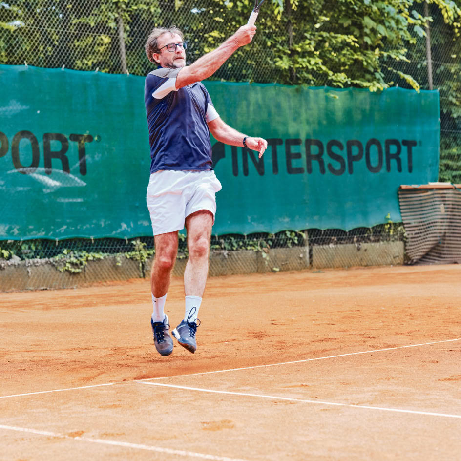 Ein Tennisspieler in blauer Kleidung und wei&szlig;en Shorts springt, um einen Ball auf einem Sandplatz zu schlagen, mit einem gr&uuml;nen Zaun im Hintergrund.