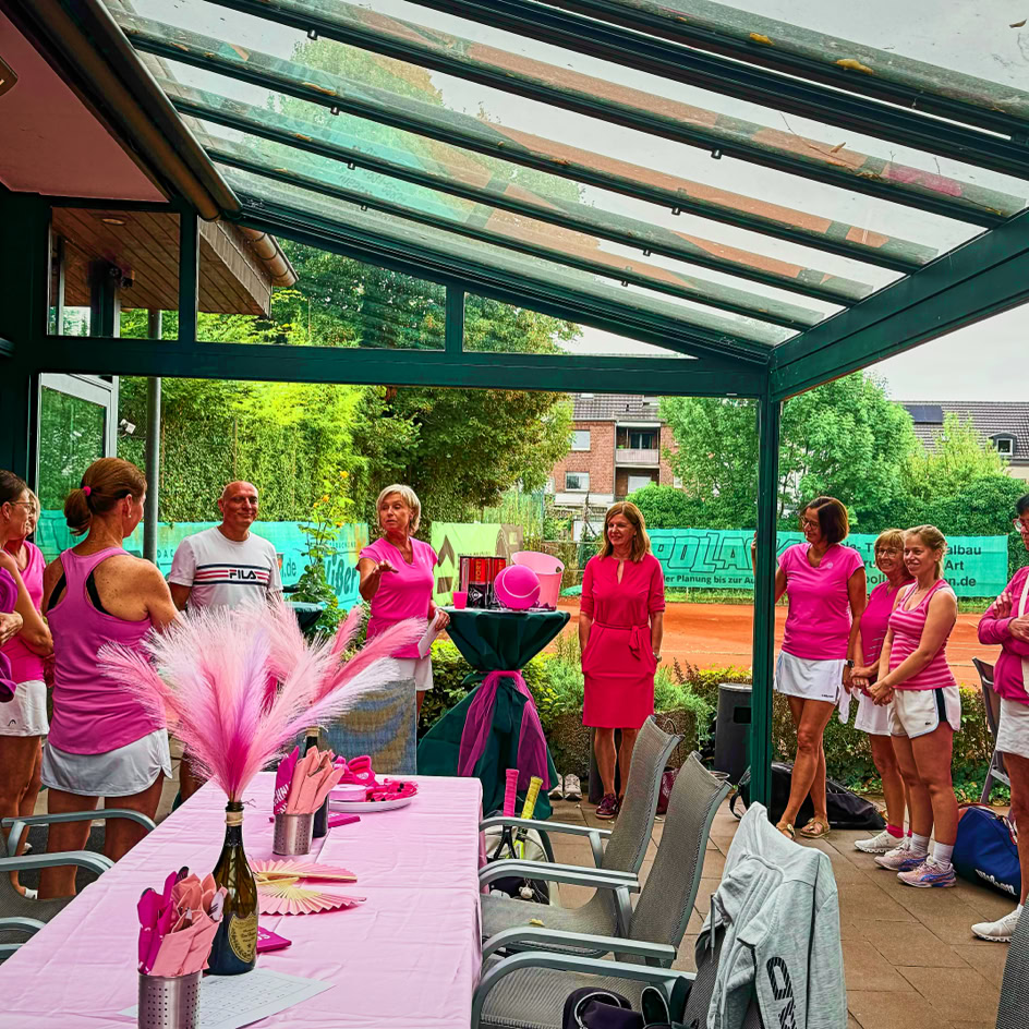 Eine Gruppe von Menschen in pinker Kleidung steht in einem &uuml;berdachten Au&szlig;enbereich, um einen Tisch mit rosa Dekorationen. Im Hintergrund sind B&auml;ume und ein Tennisplatz zu sehen.