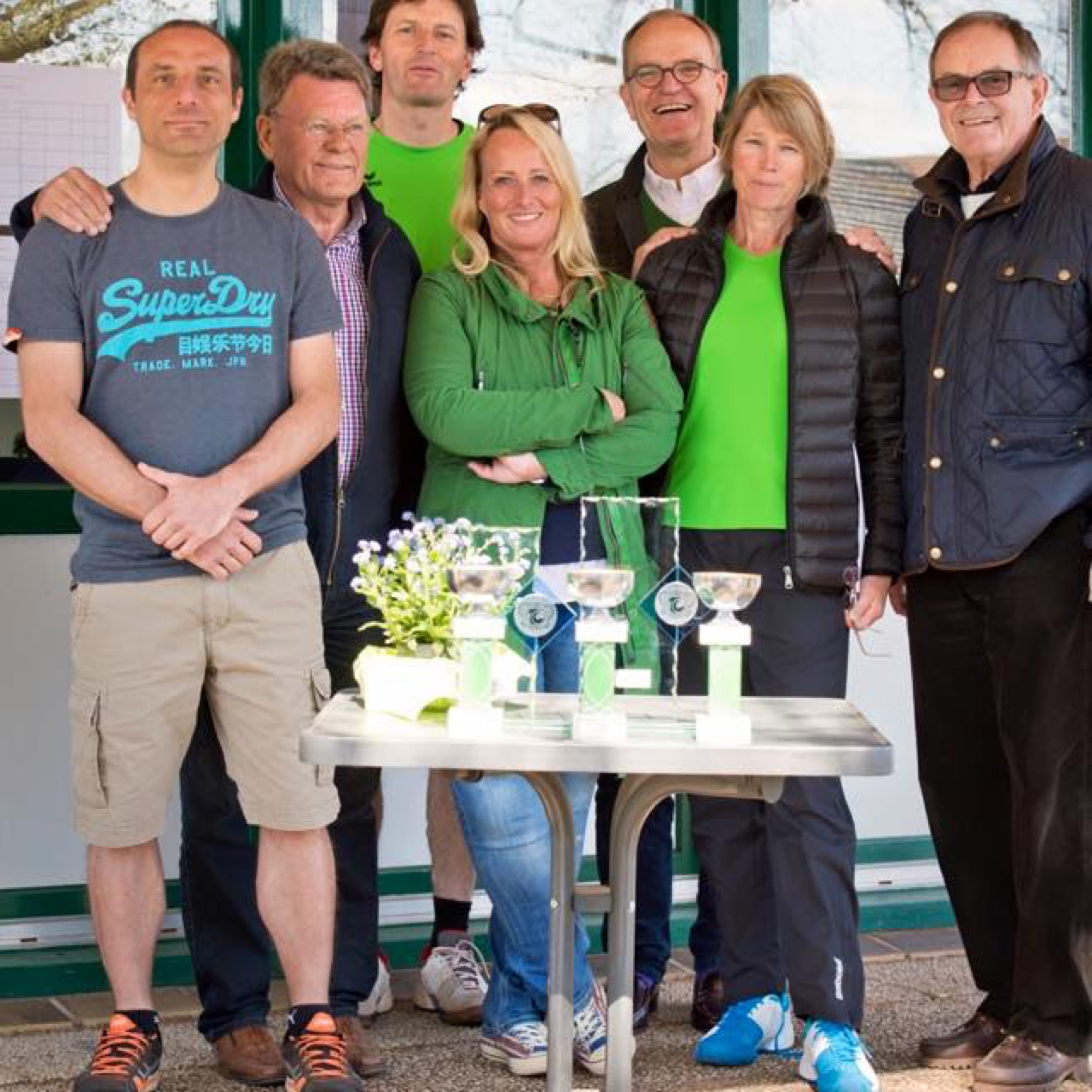 Eine Gruppe von sieben Personen steht vor einem Geb&auml;ude mit gr&uuml;nen Fenstern. Vor ihnen steht ein Tisch mit Blumen und zwei Pokalen.