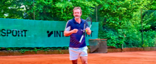 Eine Person spielt Tennis auf einem Sandplatz, umgeben von gr&uuml;nen B&auml;umen und einem Zaun mit einem Werbebanner.
