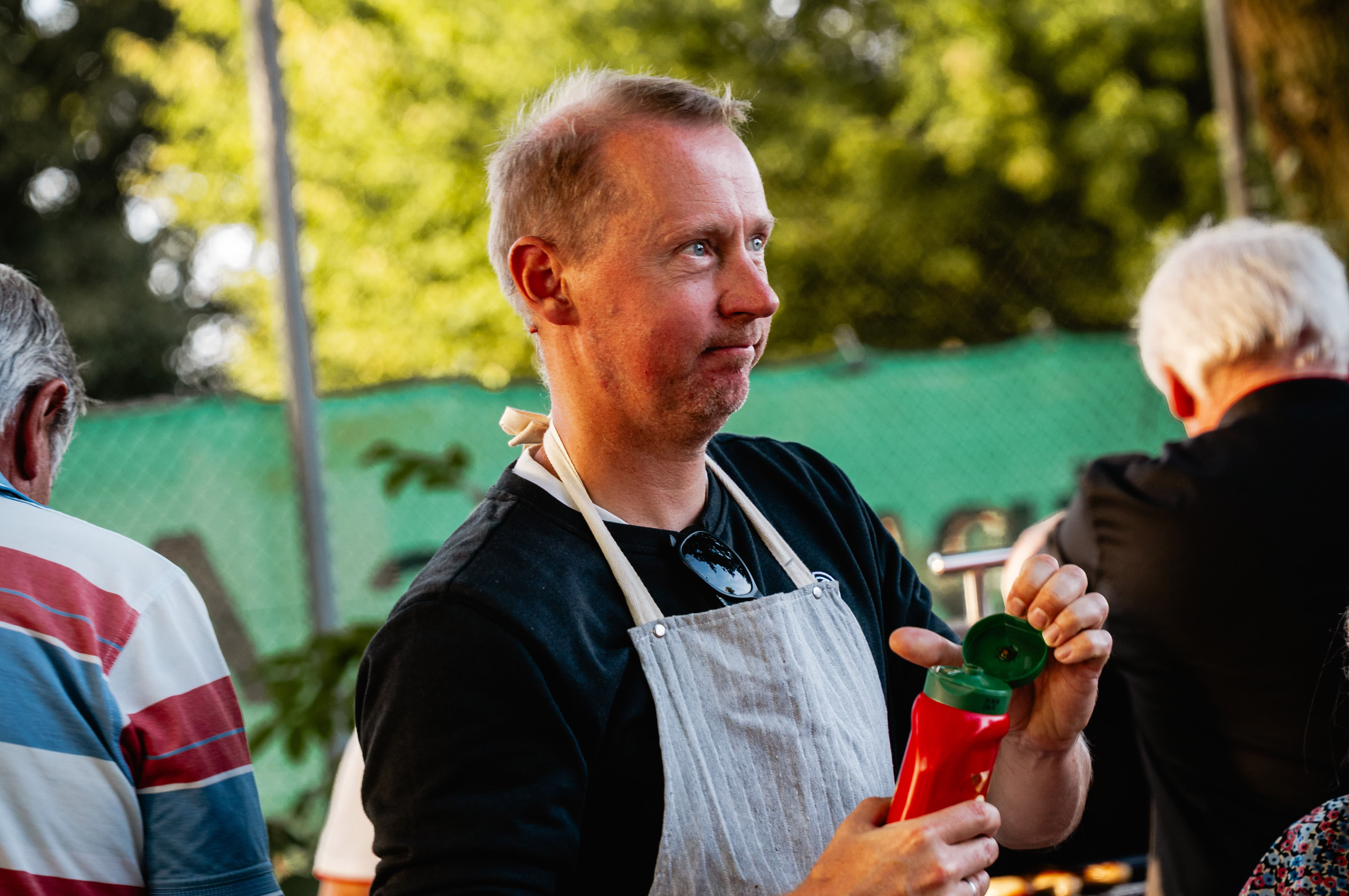 Mann in Sch&uuml;rze h&auml;lt eine rote Flasche, im Freien bei einer Veranstaltung mit anderen Menschen im Hintergrund.