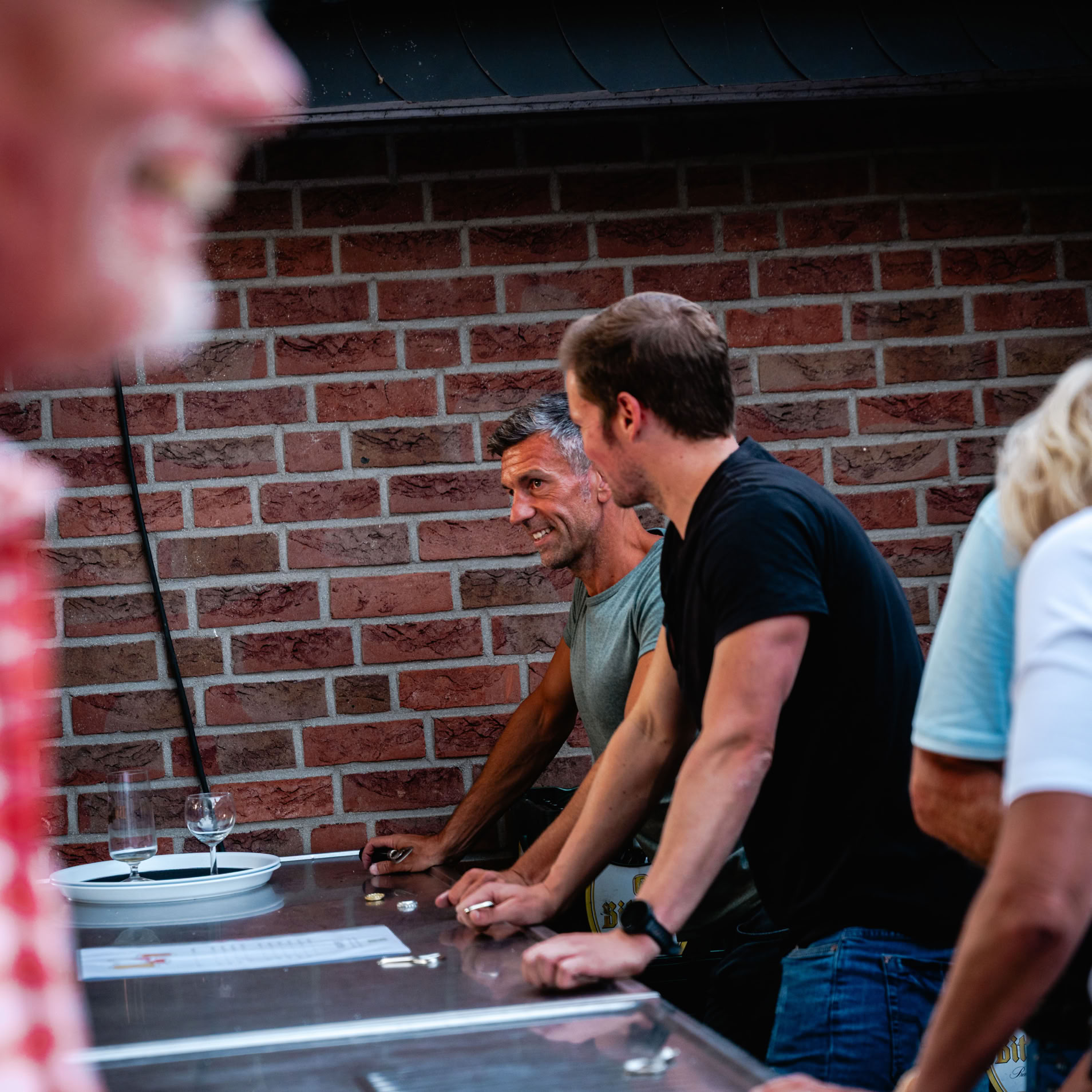 Mehrere Personen stehen an einem Tisch im Freien vor einer Backsteinmauer. Eine Person im Vordergrund tr&auml;gt ein rot-wei&szlig; kariertes Hemd.