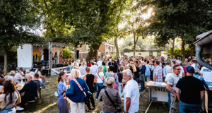 Eine gro&szlig;e Menschenmenge steht im Freien unter B&auml;umen, einige Personen sitzen an Tischen, im Hintergrund ist eine B&uuml;hne zu sehen.