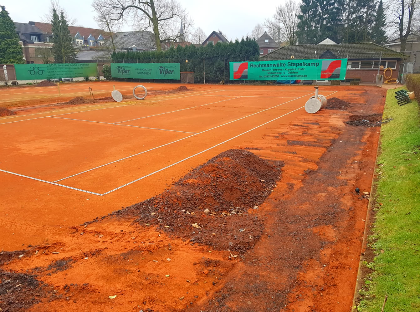 Tennisplatz mit rotem Sandbelag, umgeben von B&auml;umen und Geb&auml;uden im Hintergrund.