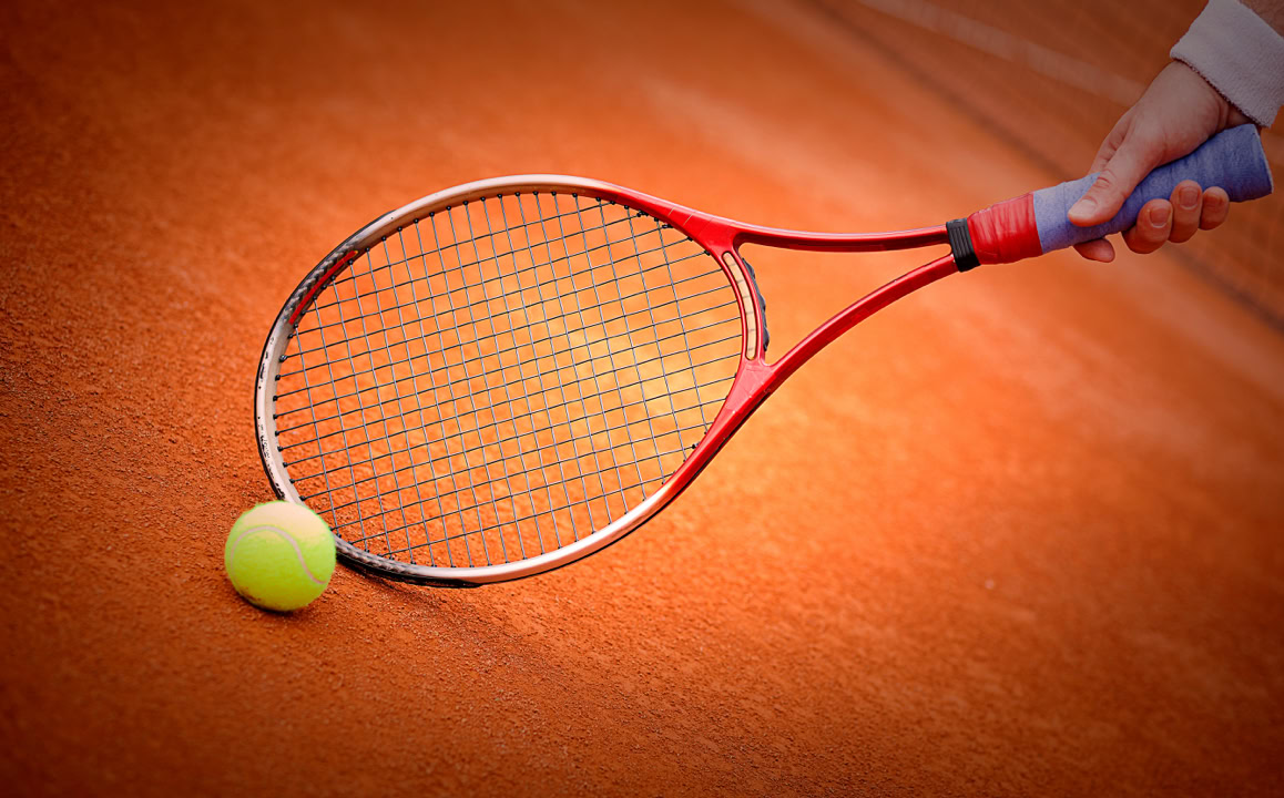 Tennisball und Tennisschl&auml;ger auf einem Sandplatz, Hand h&auml;lt den Schl&auml;ger.