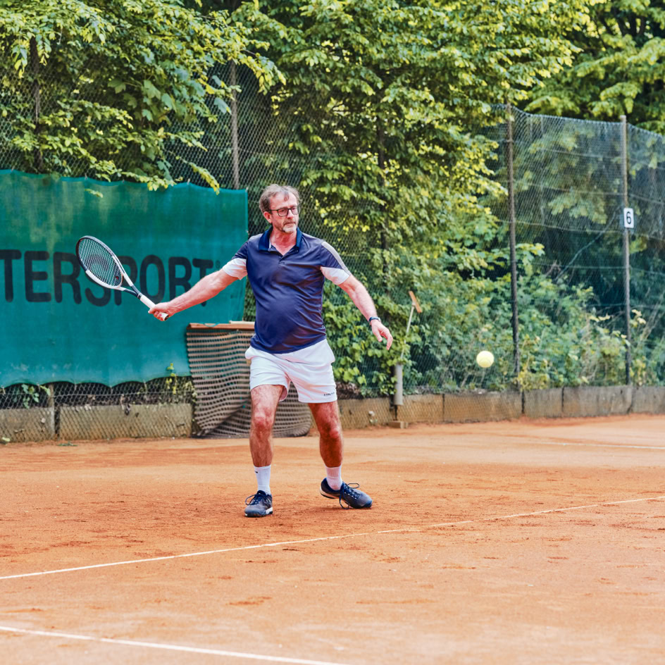 Person spielt Tennis auf einem Sandplatz, umgeben von B&auml;umen und einem gr&uuml;nen Zaun.
