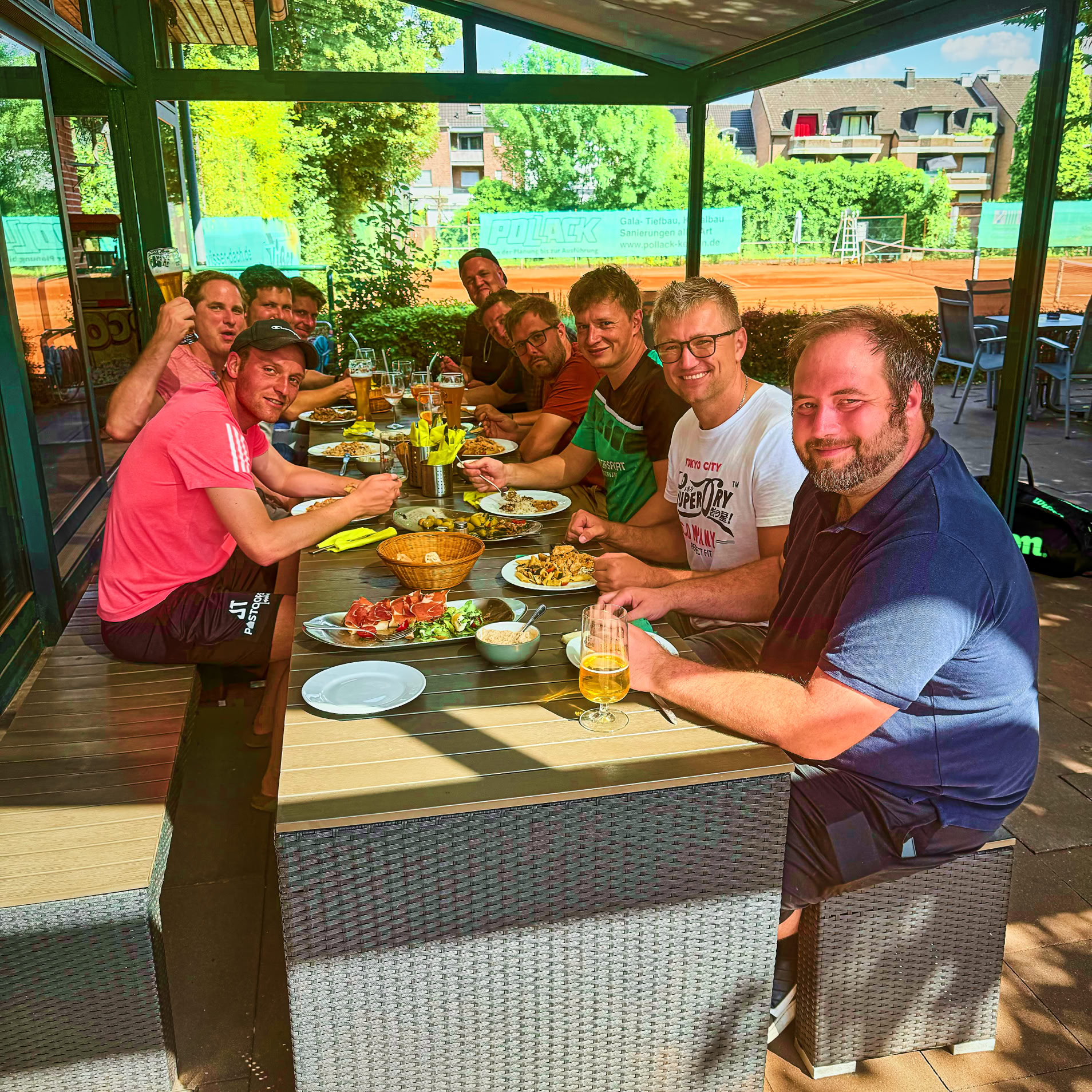 Eine Gruppe von acht Personen sitzt an einem langen Tisch im Freien und isst gemeinsam. Auf dem Tisch stehen verschiedene Speisen und Getr&auml;nke. Im Hintergrund sind B&auml;ume und Geb&auml;ude zu sehen.