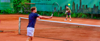 Zwei Personen spielen Tennis auf einem Sandplatz, umgeben von gr&uuml;ner Vegetation und einer Wand mit Stadtbild-Graffiti im Hintergrund.