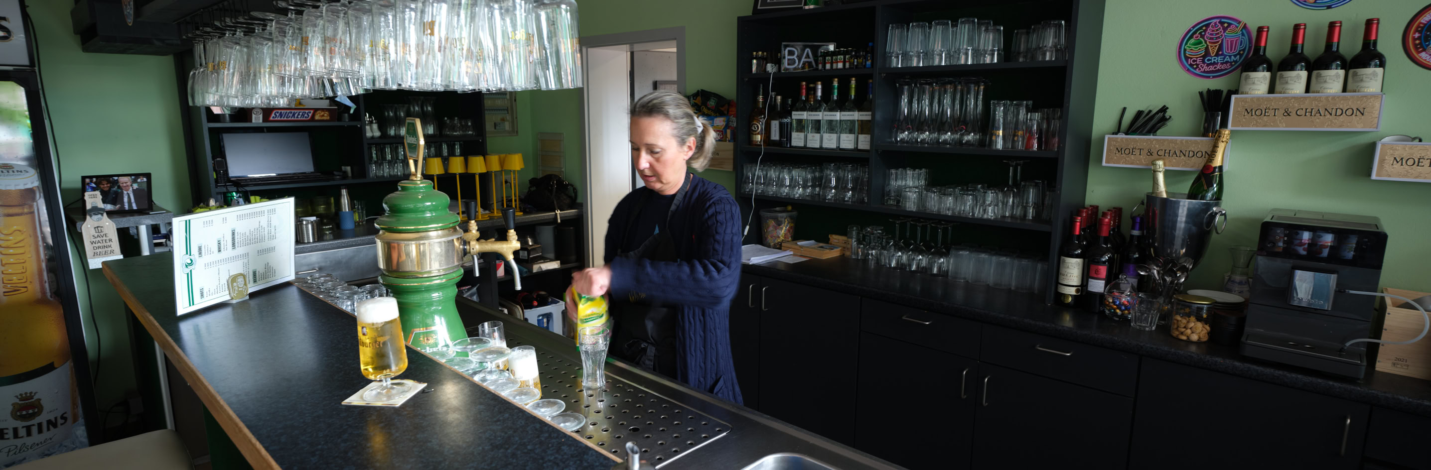 Eine Person steht hinter einer Bar mit gr&uuml;nen W&auml;nden, umgeben von Gl&auml;sern und Flaschen.