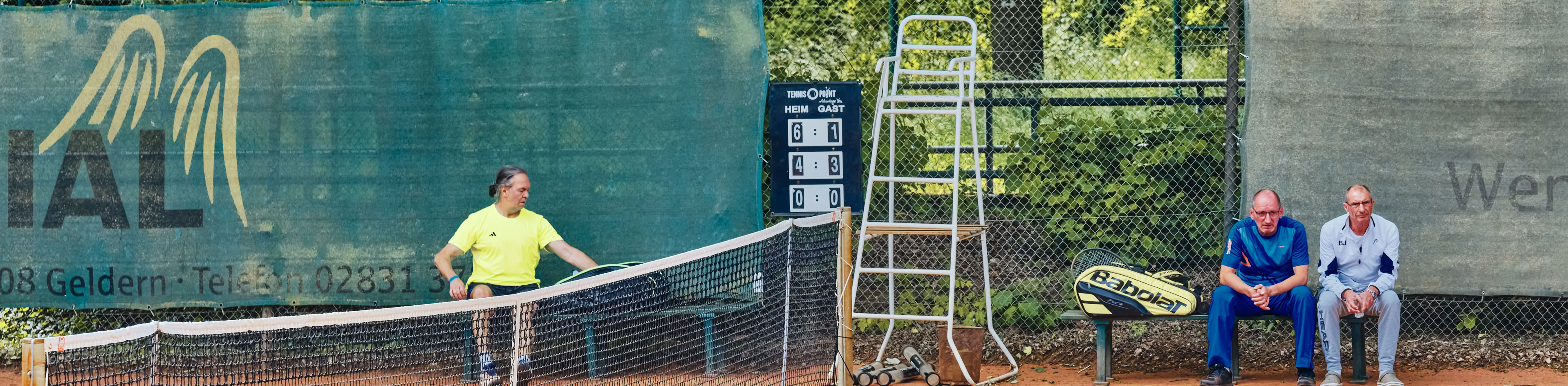 Ein Tennisplatz mit einem Spieler in gelbem Shirt, der den Ball schl&auml;gt, und zwei Personen, die auf einer Bank am Rand sitzen. Im Hintergrund sind B&auml;ume und ein Zaun zu sehen.