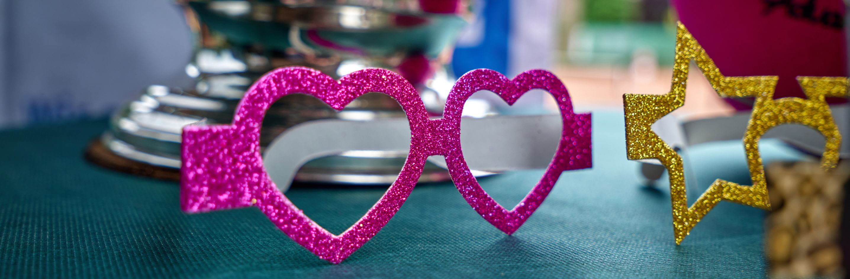 Rosa herzf&ouml;rmige Brille und goldene sternf&ouml;rmige Brille auf einem Tisch vor einem silbernen Pokal.