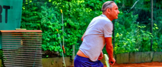 Ein Mann in Sportkleidung steht auf einem Tennisplatz mit rotem Sand, im Hintergrund sind gr&uuml;ne B&auml;ume und ein Teil eines Schildes mit der Aufschrift "SPORT" zu sehen.