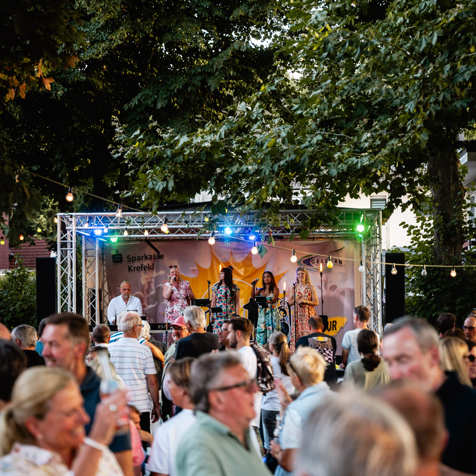 Menschenmenge bei einem Freiluftkonzert, B&uuml;hne mit Band im Hintergrund, B&auml;ume und Lichterketten.