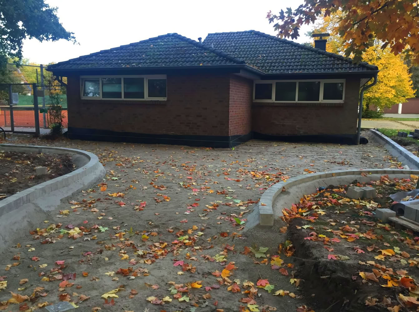 Backsteinhaus mit dunklem Dach, umgeben von herbstlichen B&auml;umen und Laub auf dem Boden.
