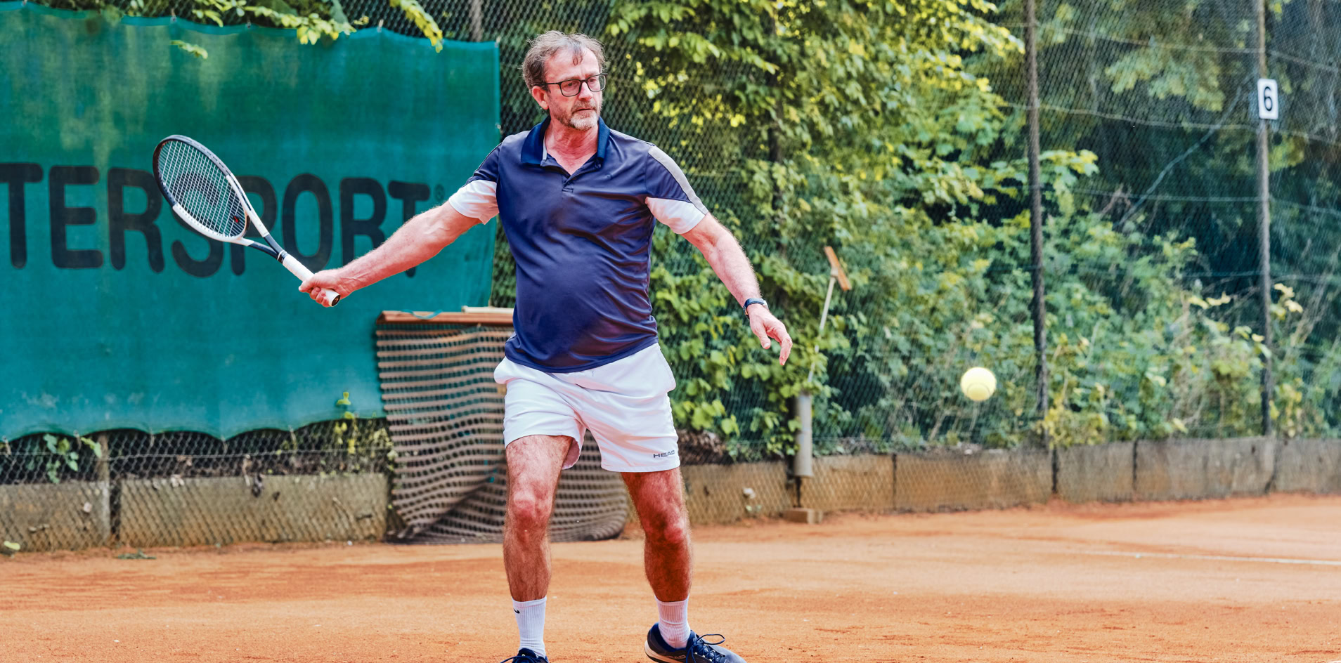 Person spielt Tennis auf einem Sandplatz, umgeben von B&auml;umen und einem gr&uuml;nen Zaun.