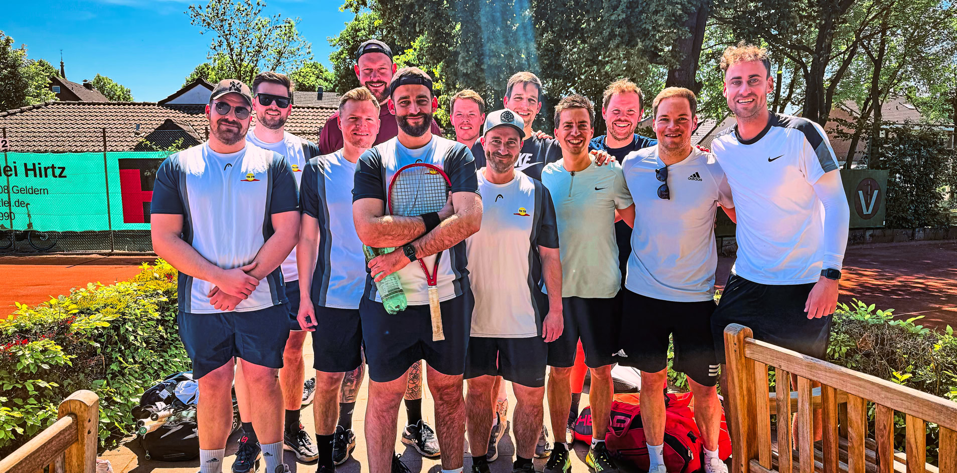 Eine Gruppe von Menschen in Sportkleidung steht auf einem Tennisplatz, umgeben von B&auml;umen und einem Geb&auml;ude im Hintergrund.