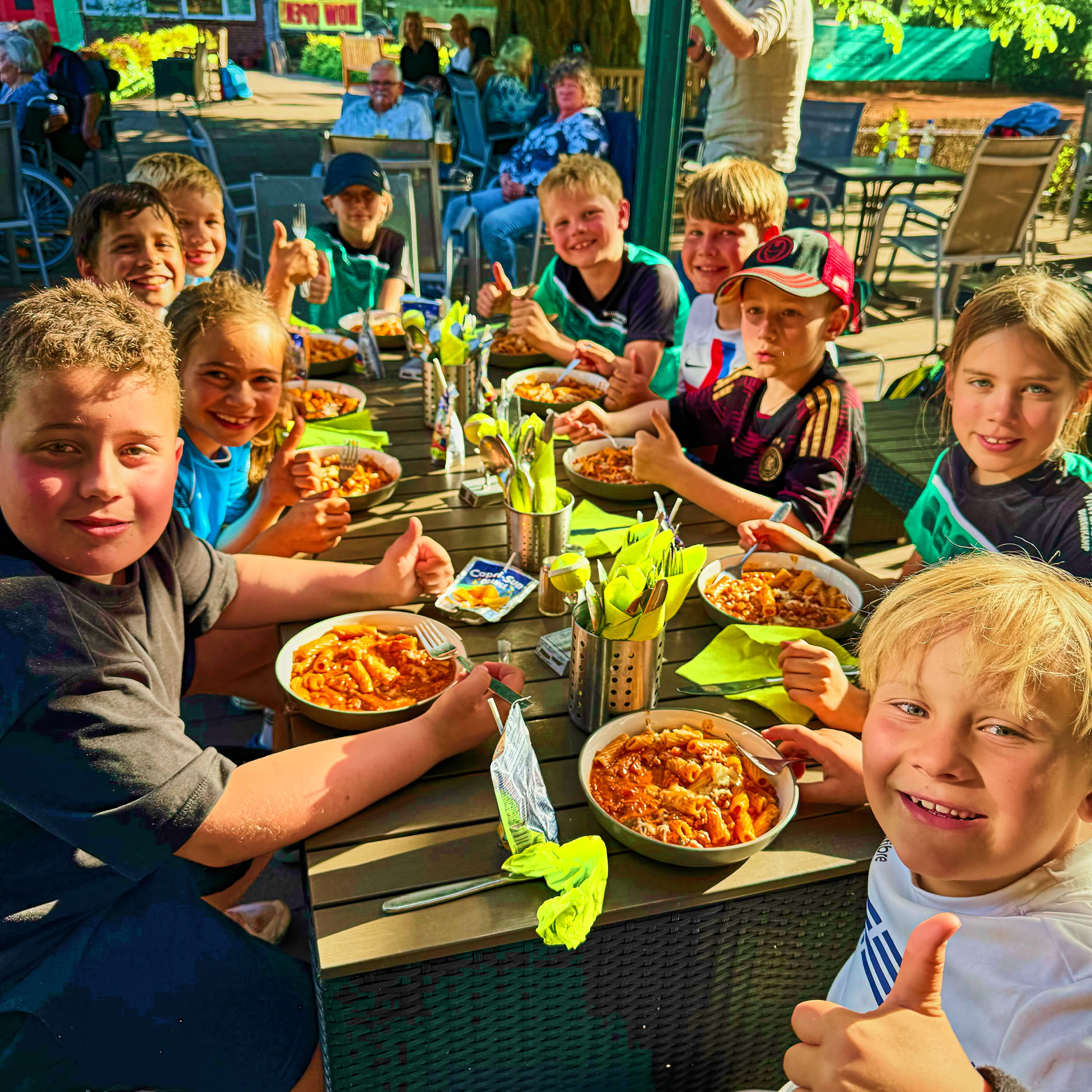 Gruppe von Kindern sitzt an einem Tisch im Freien und isst Pasta, w&auml;hrend eines der Kinder den Daumen hoch zeigt.
