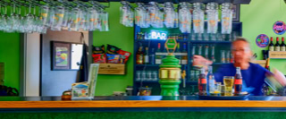 Eine Person steht hinter einer Bar mit gr&uuml;ner Wand, Gl&auml;sern und Flaschen im Hintergrund.