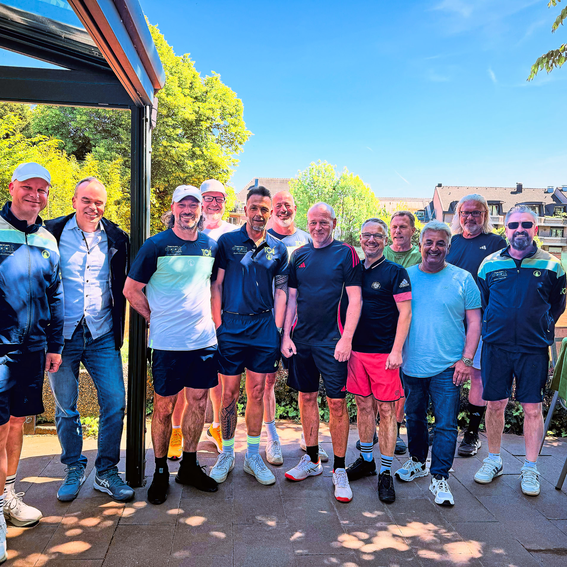 Eine Gruppe von elf Personen steht drau&szlig;en auf einer Terrasse, umgeben von B&auml;umen und einem klaren Himmel. Einige tragen Sportkleidung.