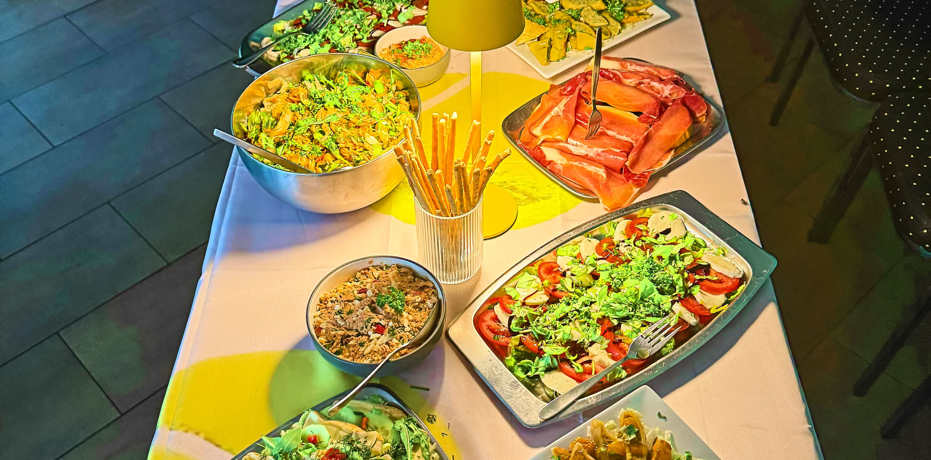 Ein langer Tisch mit verschiedenen Salaten, Brotstangen, und anderen Speisen, umgeben von St&uuml;hlen.