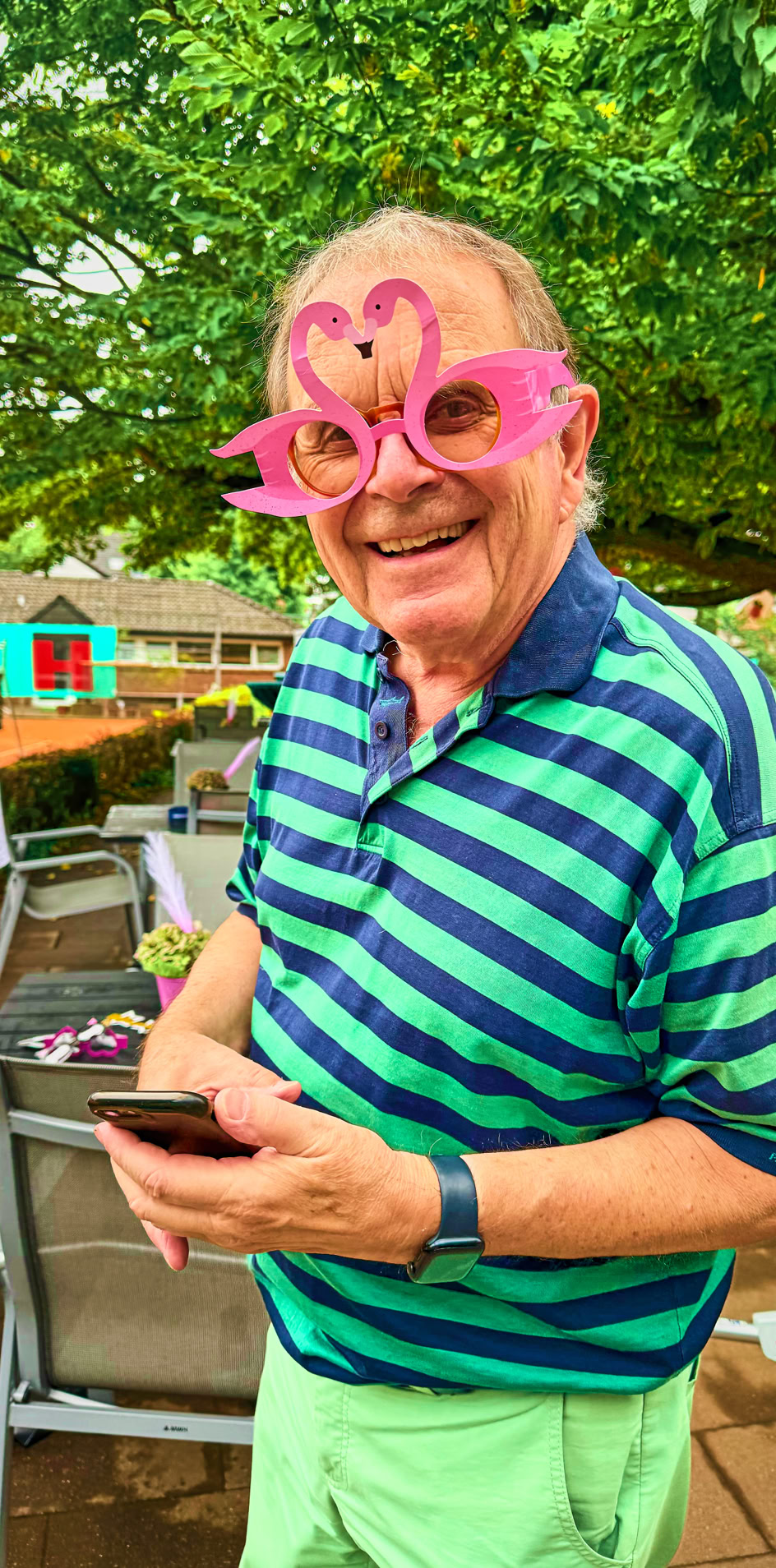 &Auml;lterer Mann mit gestreiftem Polohemd und pinker Flamingo-Brille l&auml;chelt, w&auml;hrend er ein Smartphone h&auml;lt; im Hintergrund ein weiterer Mann und Gartenm&ouml;bel unter einem Baum.
