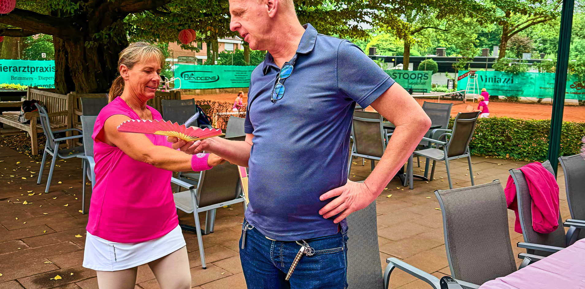 Ein Mann und eine Frau tanzen auf einer Terrasse unter einem Glasdach; im Vordergrund ein Tisch mit rosa Tischdecke und Dekoration.