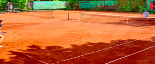 Zwei Personen spielen Tennis auf einem Sandplatz, umgeben von gr&uuml;nen B&auml;umen und Werbebannern.