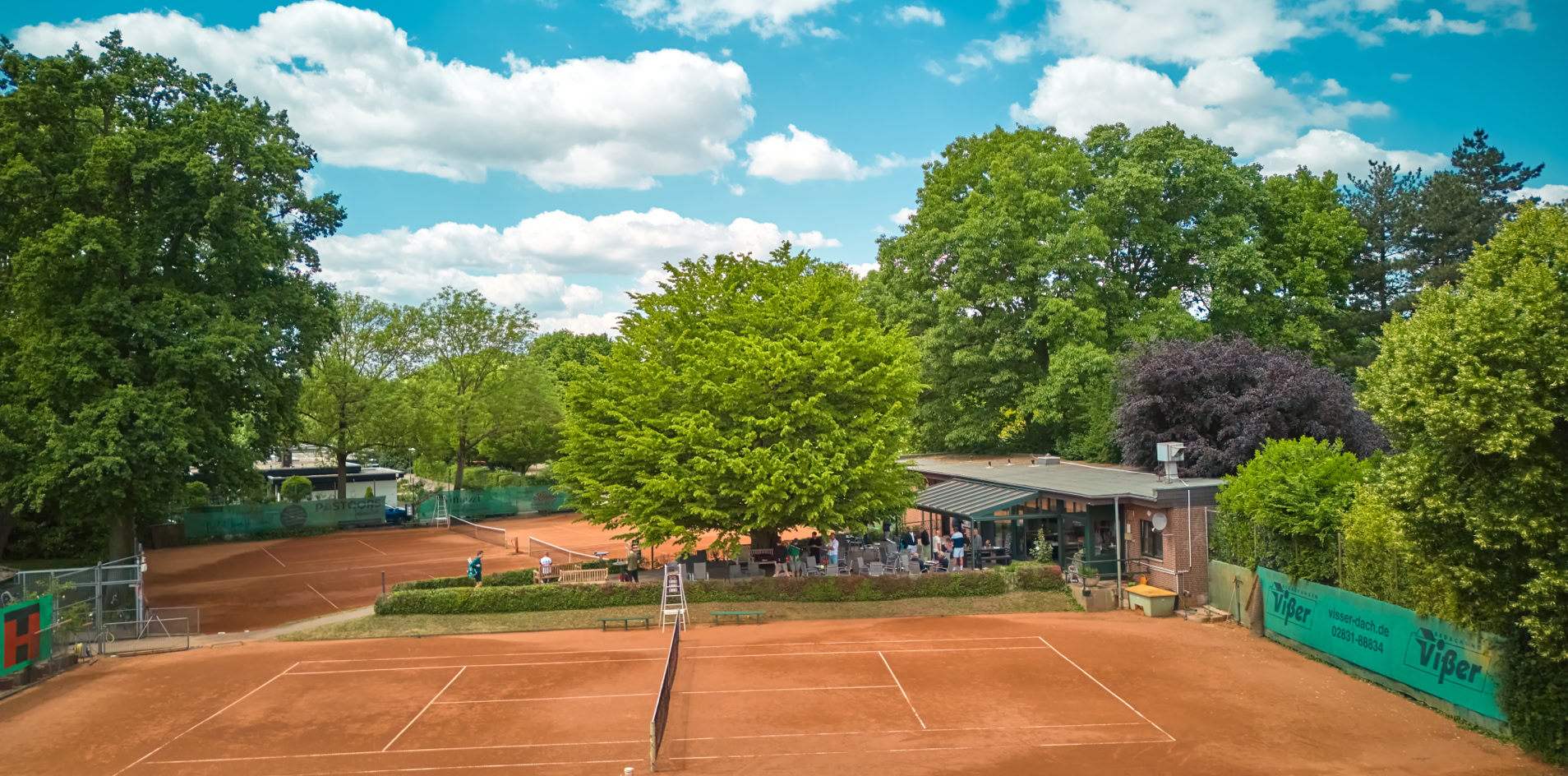 Zwei Sandtennispl&auml;tze umgeben von B&auml;umen, mit einem Geb&auml;ude im Hintergrund unter blauem Himmel mit Wolken.