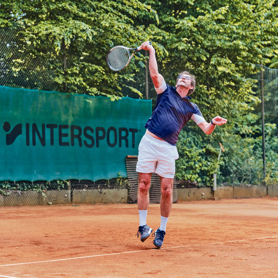 Ein Tennisspieler in blauer Kleidung und wei&szlig;en Shorts schl&auml;gt auf einem Sandplatz einen Ball auf, im Hintergrund sind gr&uuml;ne B&auml;ume und ein Werbebanner zu sehen.