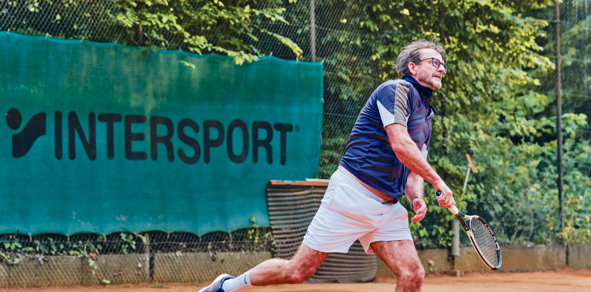 Ein Mann spielt Tennis auf einem Sandplatz, umgeben von B&auml;umen, mit einem "Intersport"-Banner im Hintergrund.