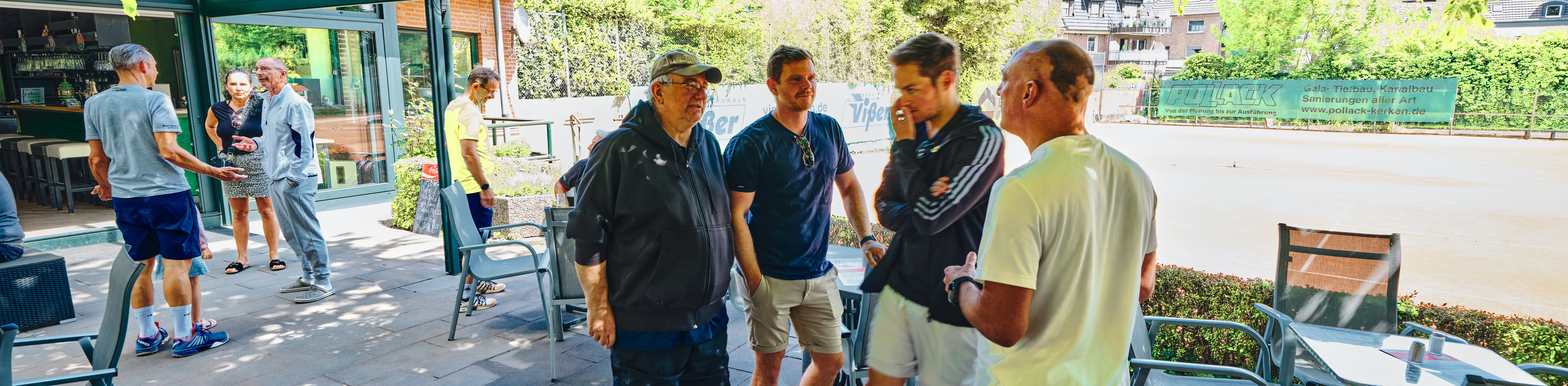 Mehrere Personen stehen und unterhalten sich auf einer Terrasse mit Tischen und St&uuml;hlen, umgeben von B&auml;umen und Pflanzen.