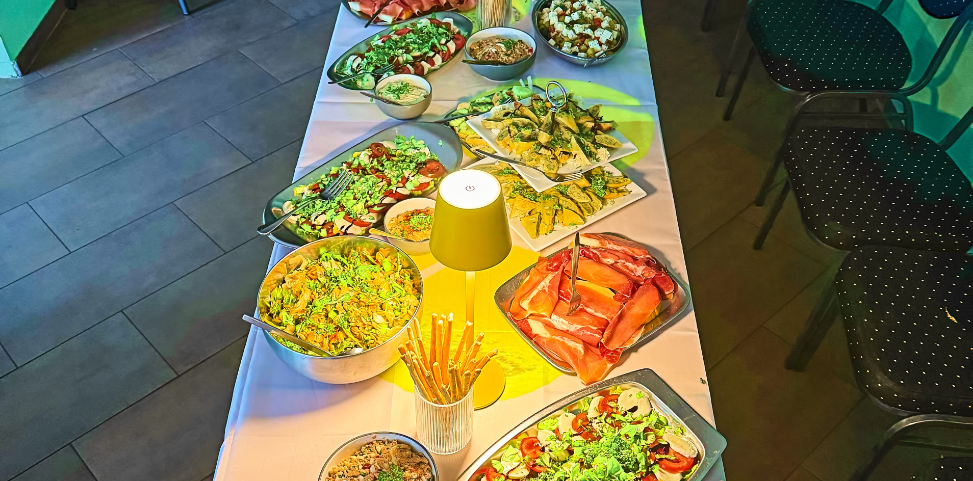 Ein langer Tisch mit verschiedenen Sch&uuml;sseln voller Salate und Speisen, umgeben von St&uuml;hlen in einem Raum mit gr&uuml;nen W&auml;nden und Kerzen.