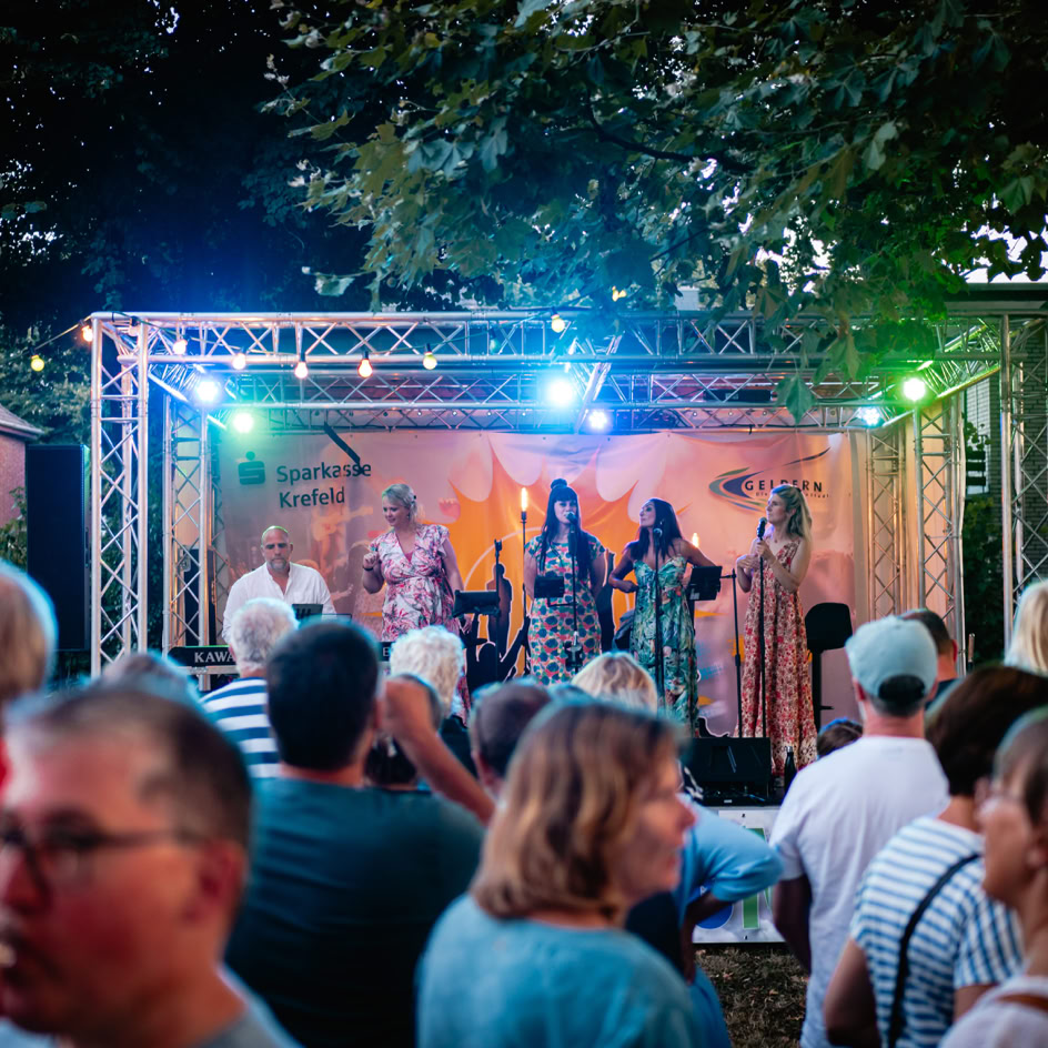 Eine Gruppe von Menschen steht vor einer B&uuml;hne im Freien, auf der mehrere Personen unter bunten Lichtern auftreten.