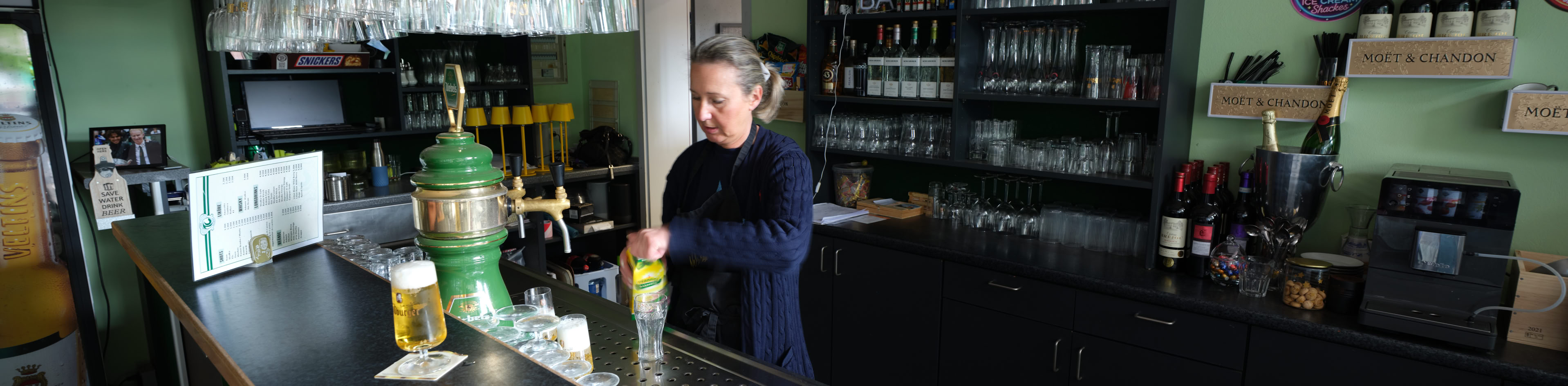 Eine Person steht hinter einer Bar mit gr&uuml;nen W&auml;nden, umgeben von Gl&auml;sern und Flaschen.