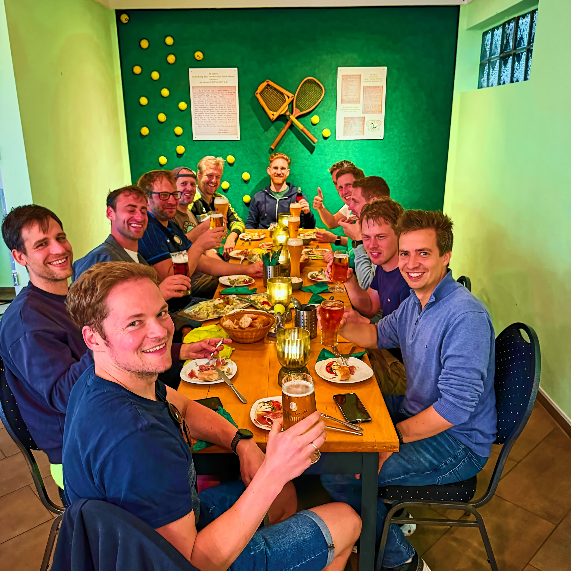 Eine Gruppe von Personen sitzt an einem Tisch in einem Raum mit gr&uuml;ner Wand, auf der Tennisschl&auml;ger und B&auml;lle dekoriert sind.