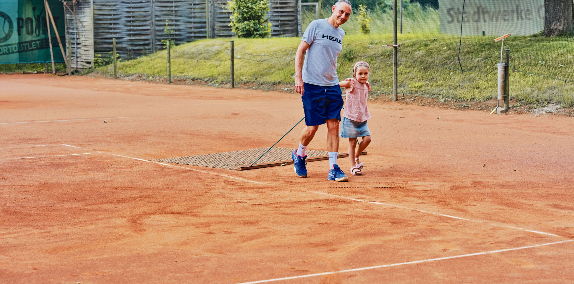 Ein Mann und ein Kind ziehen ein Netz &uuml;ber einen Sandtennisplatz.