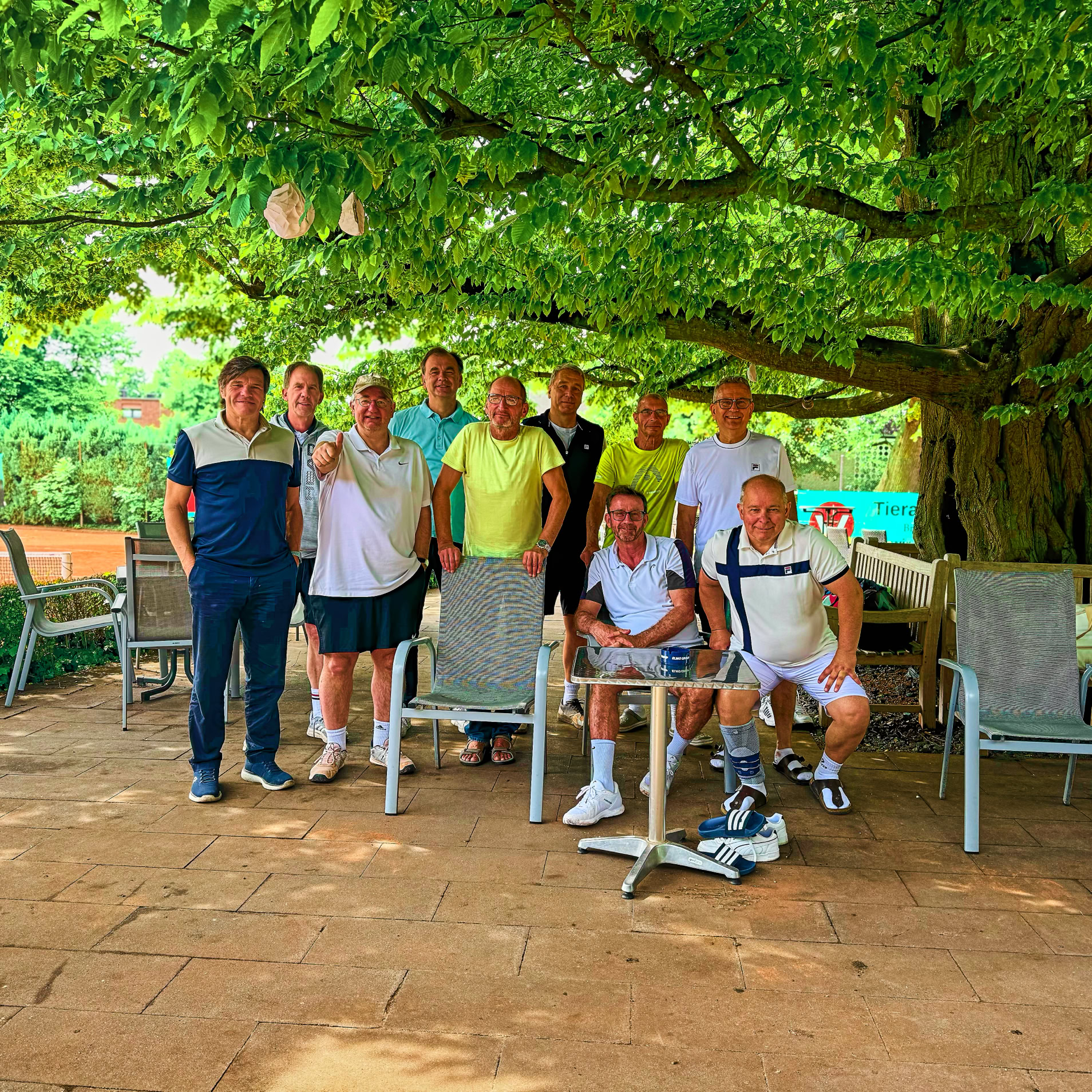 Eine Gruppe von neun Personen steht und sitzt unter einem gro&szlig;en Baum mit dichtem Laub, umgeben von Gartenm&ouml;beln auf einer Terrasse. Im Hintergrund ist ein Tennisplatz zu sehen.