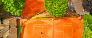 Luftaufnahme von zwei Tennispl&auml;tzen mit rotem Sand, umgeben von gr&uuml;nen B&auml;umen und Geb&auml;uden.