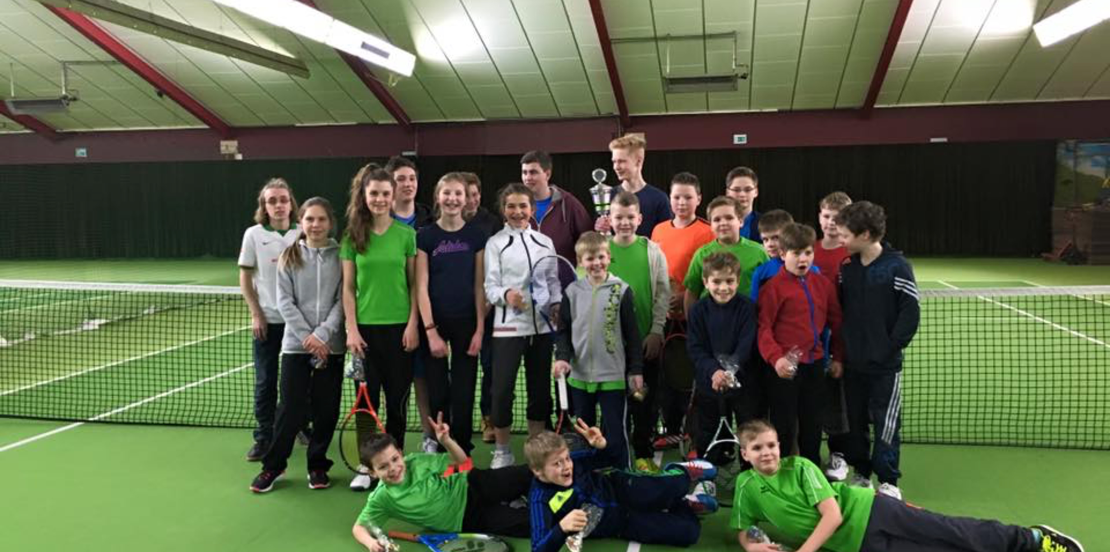 Gruppe von Personen steht und liegt auf einem Tennisplatz in einer Halle.