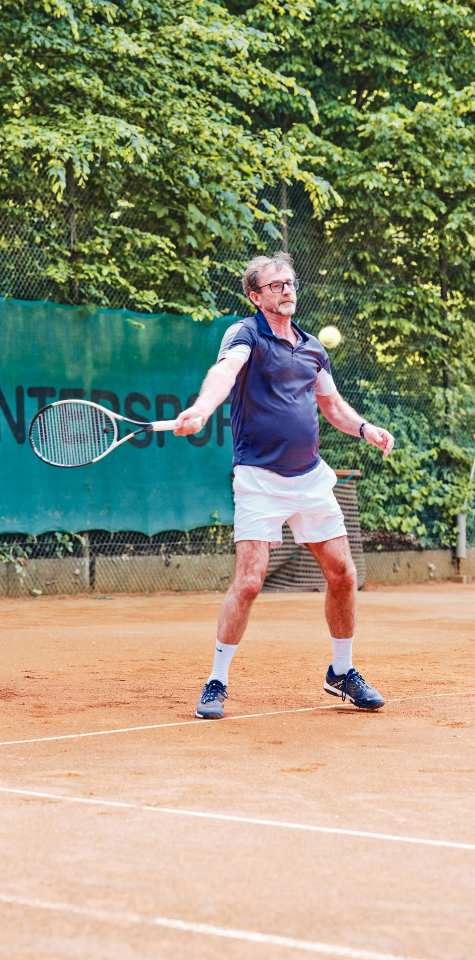 Eine Person spielt Tennis auf einem Sandplatz, umgeben von B&auml;umen, mit einem gr&uuml;nen Zaun im Hintergrund.