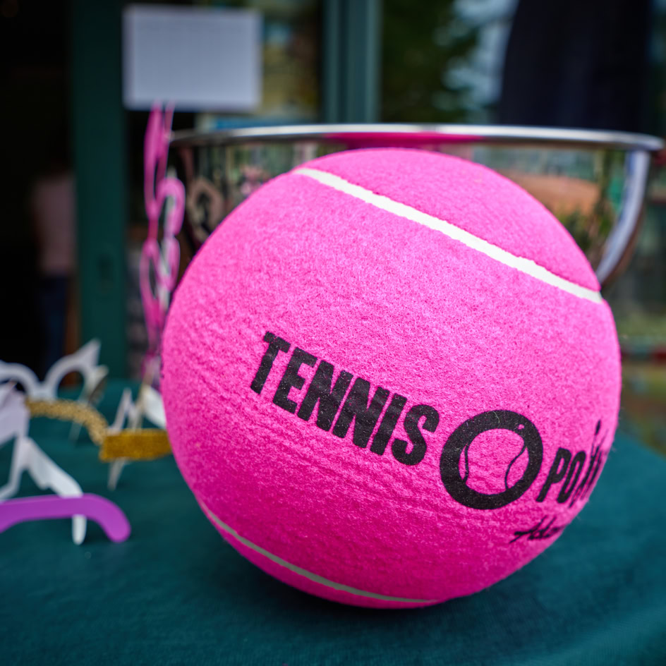 Ein gro&szlig;er pinker Tennisball mit der Aufschrift "TENNIS" liegt auf einem Tisch mit gr&uuml;nem Tuch, im Hintergrund ein silberner Pokal.