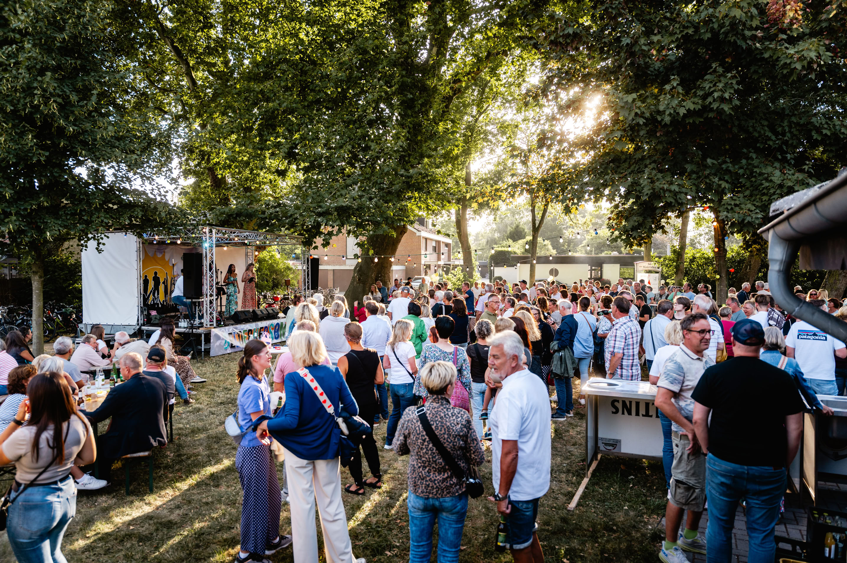 Menschenmenge bei einer Veranstaltung im Freien, umgeben von B&auml;umen, mit St&auml;nden und Tischen im Hintergrund.