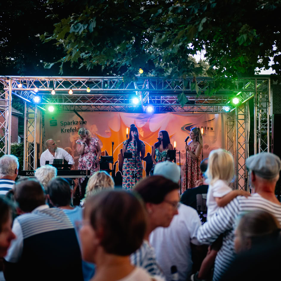 Eine Menschenmenge steht vor einer B&uuml;hne im Freien, auf der mehrere Personen unter bunten Lichtern auftreten.