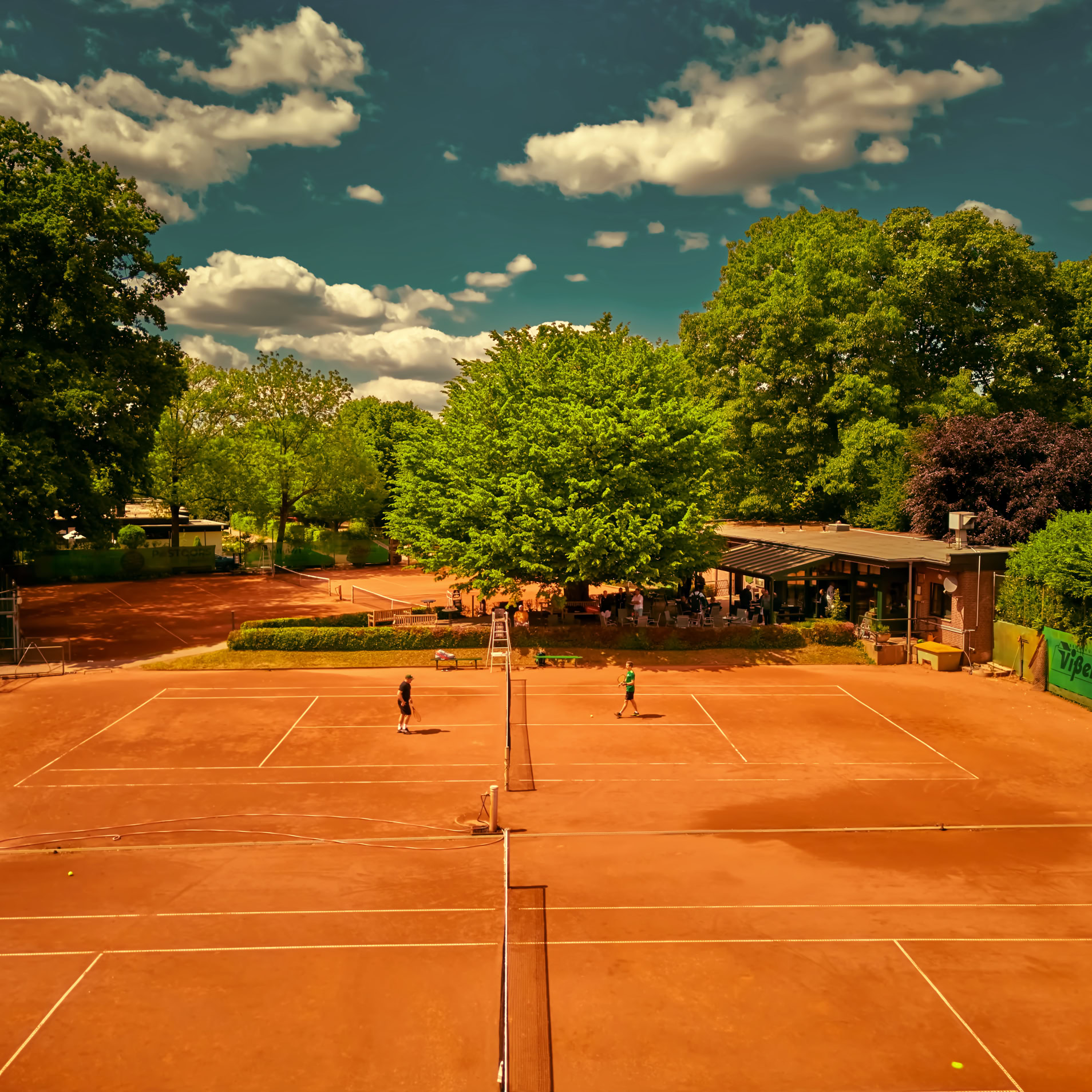 Zwei Personen spielen Tennis auf einem Sandplatz, umgeben von B&auml;umen und einem Geb&auml;ude im Hintergrund.