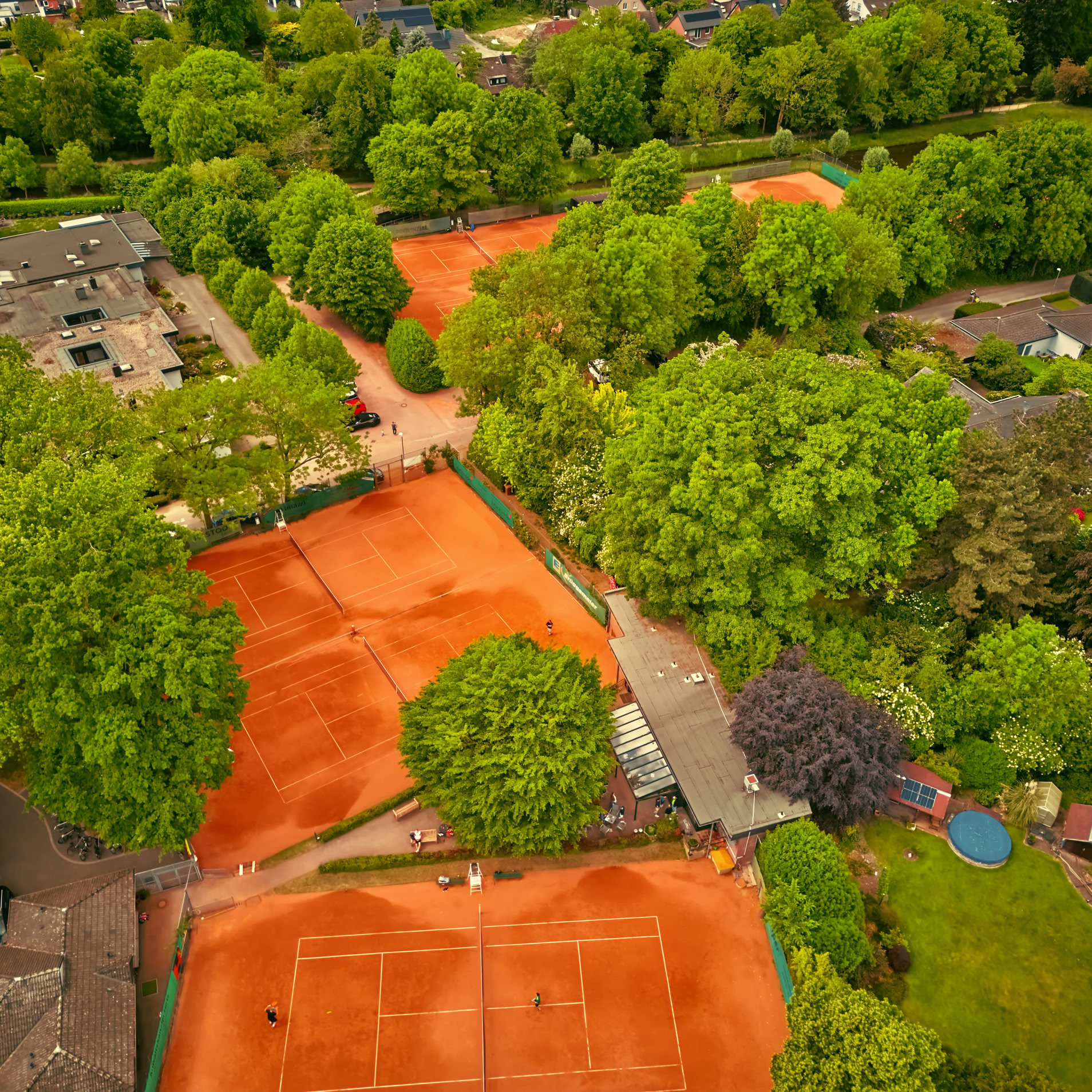 Luftaufnahme von mehreren Tennispl&auml;tzen mit rotem Sand, umgeben von gr&uuml;nen B&auml;umen und Geb&auml;uden.