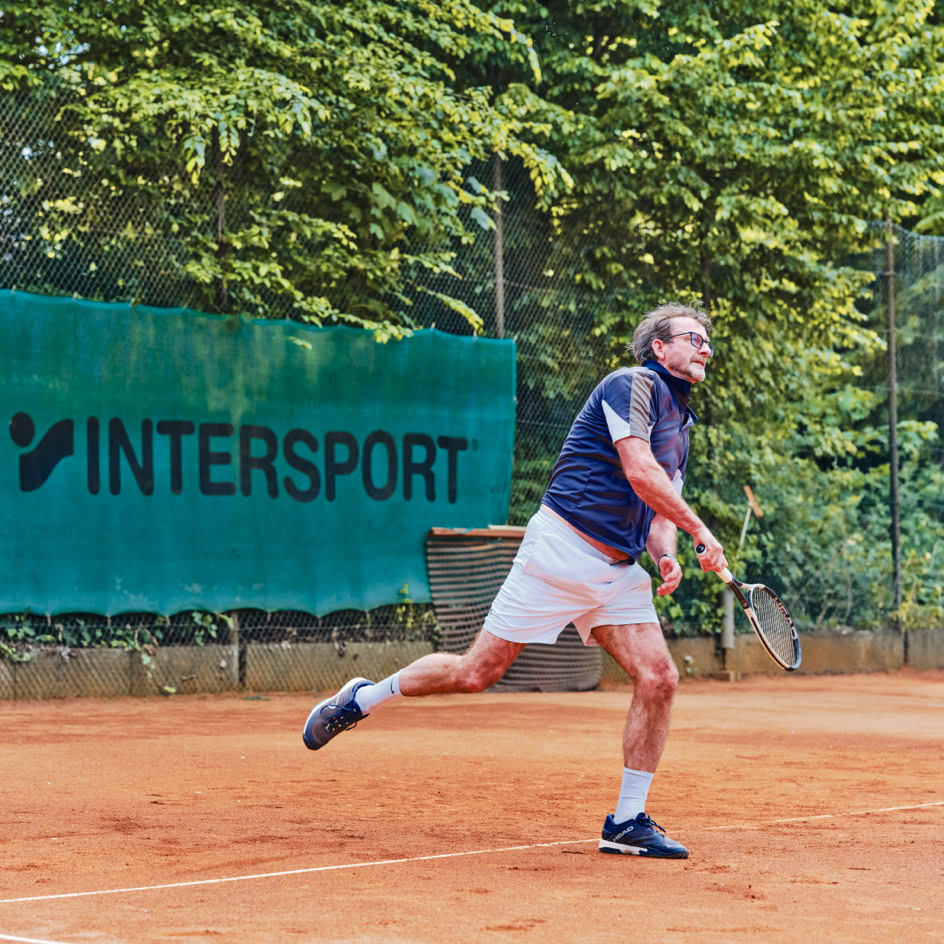 Ein Mann spielt Tennis auf einem Sandplatz, umgeben von B&auml;umen, mit einem "Intersport"-Banner im Hintergrund.