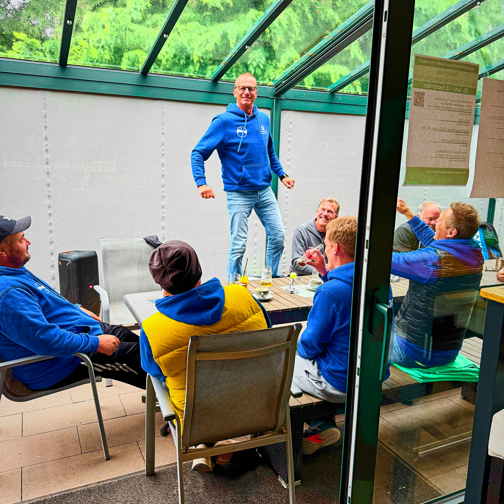 Eine Gruppe von Menschen sitzt um einen Tisch in einem Raum mit Glasdach, w&auml;hrend eine Person auf dem Tisch steht und spricht.
